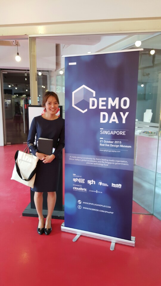 Woman in blue dress holding white bag and standing next to a Demo Day banner