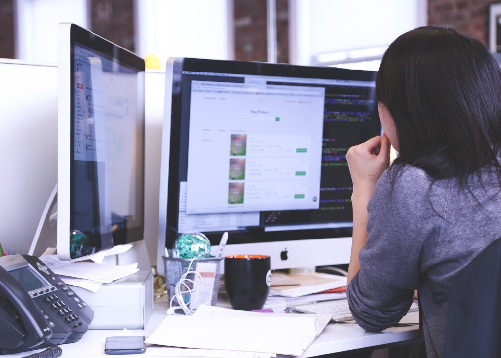 Back of woman sitting at office desk looking at dual computer screens. 